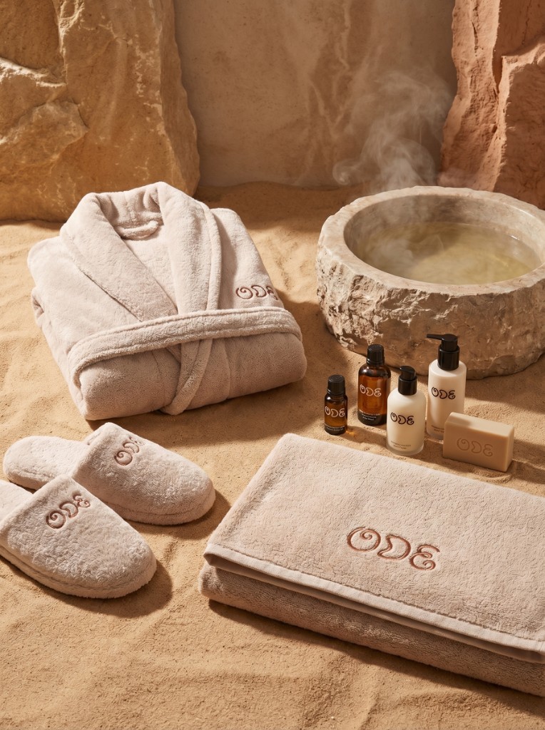 Ode spa suite: robe, slippers, towels, apothecary bottles, soap, and steaming stone basin on sand